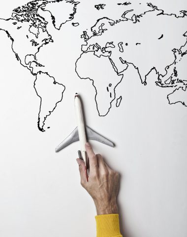 Crop person in vibrant yellow clothes holding little gray plane against contour map on white wall as concept of tourism and adventure